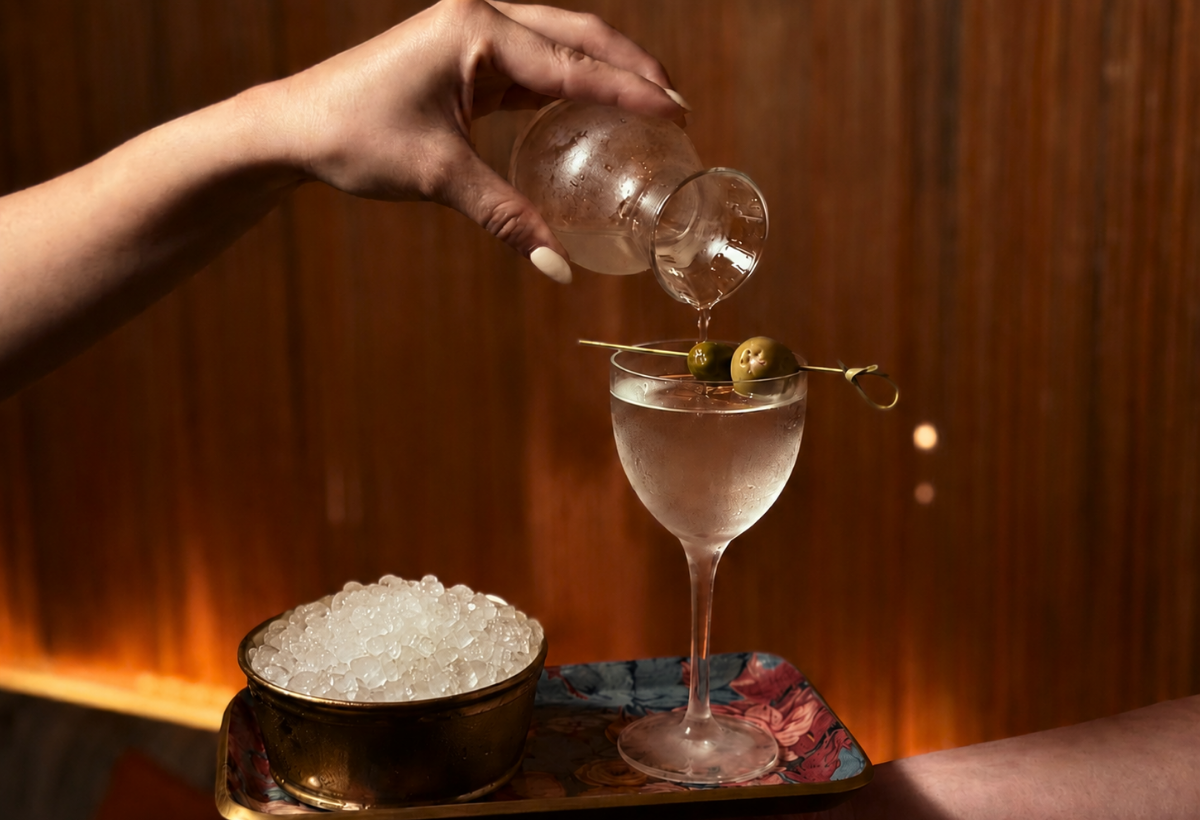 A hand pours liquid into a coupe glass with a skewer of two olives resting on the rim, ice bucket nearby on a tray.
