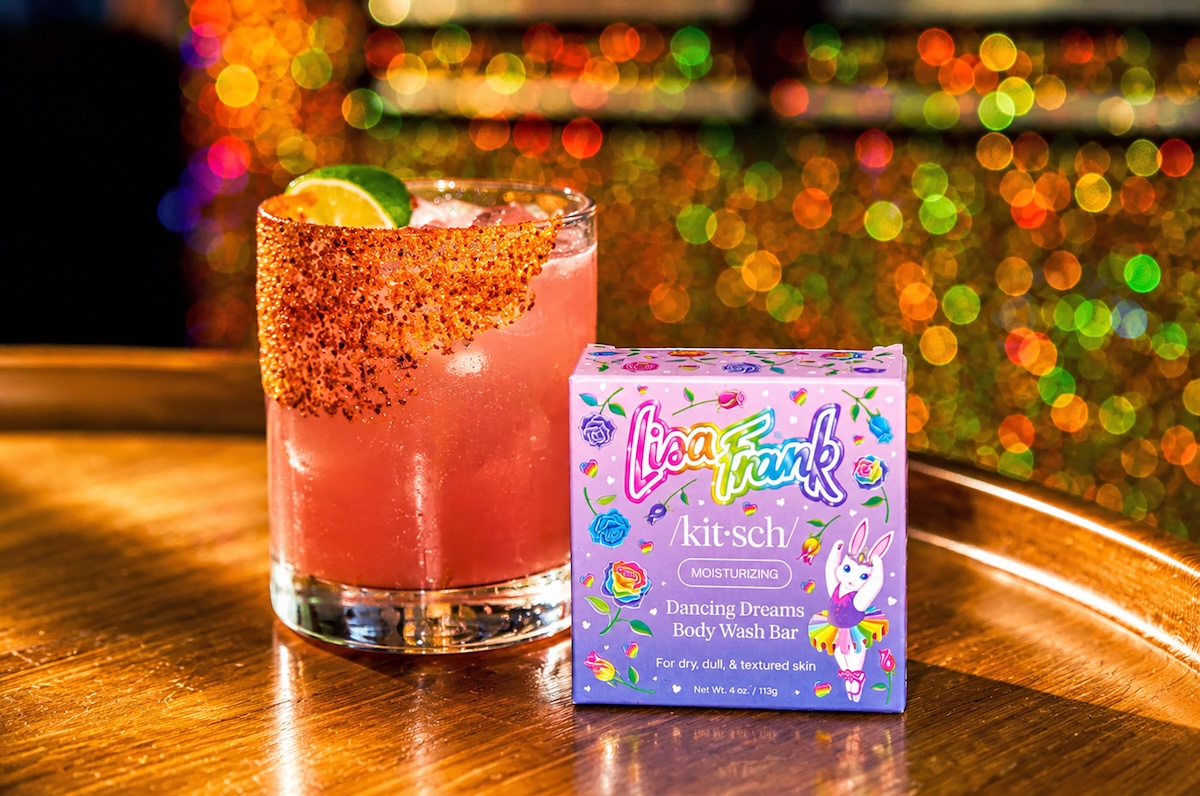 Pink cocktail with a sugared rim and lime wedge, next to a colorful box labeled 'Dancing Dreams Body Wash Bar' on a wooden bar counter.