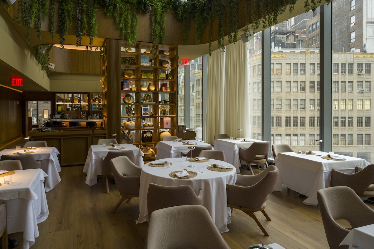 Elegant restaurant dining room with white-tablecloth tables, beige chairs, and a hanging-greenery ceiling near large windows overlooking a cityscape.