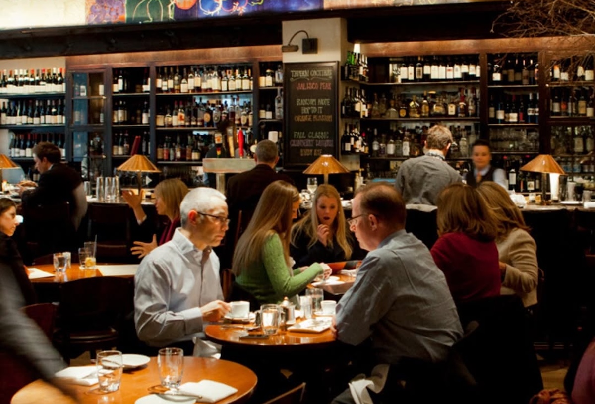 Lively restaurant scene with groups of people dining at round wooden tables and a long bar lined with bottles in the background.