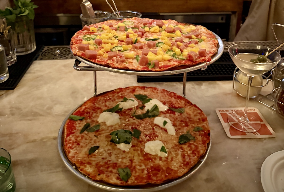 Two large pizzas on a metal stand atop a bar counter: the top pie has ham, pineapple, and veg; the bottom pie is a basil-tleck mozzarella cheese pizza.