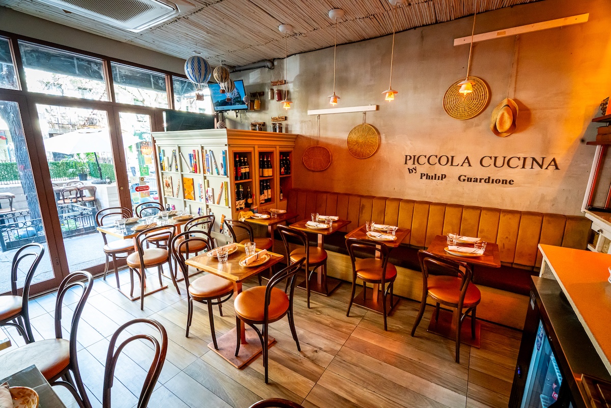 Cozy Italian restaurant interior with wooden tables and brown chairs, a long booth along the wall, warm pendant lights, and shelves of books and bottles.
