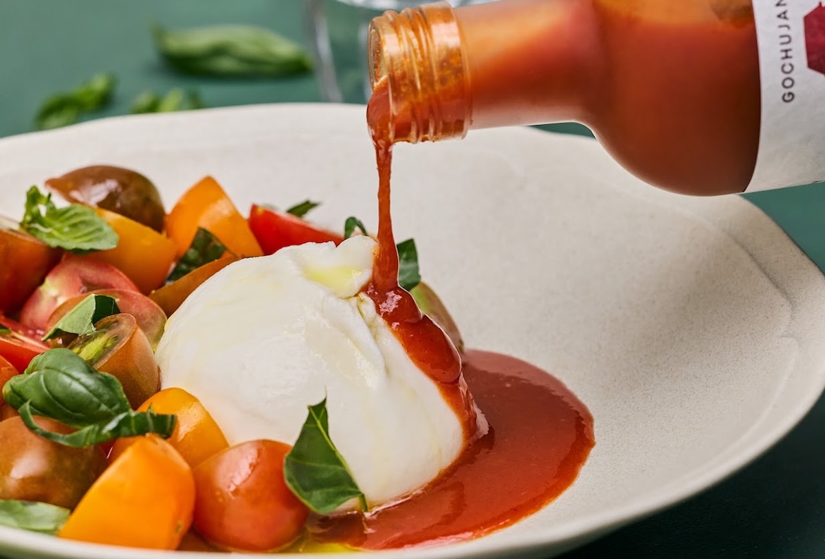 Creamy burrata on a tomato-basil salad as red sauce is poured over it from a bottle.