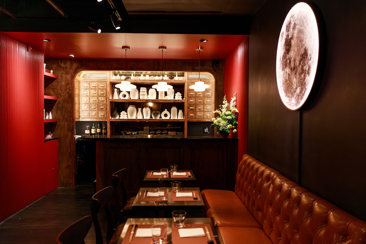 Cozy restaurant dining area with a red accent wall, leather banquette, and a bar displaying white dishes behind glass.