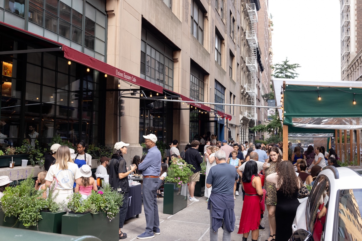 Crowd of people at an outdoor urban cafe event along a city street near a cafe awning.