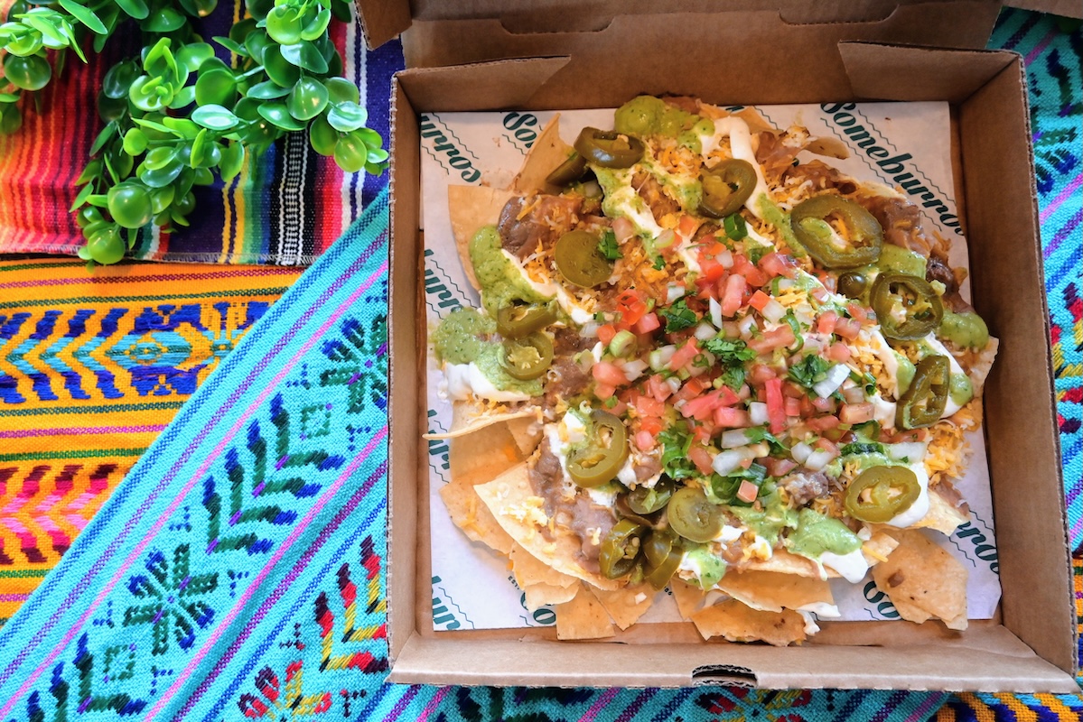 Box of loaded nachos with jalapeños, diced tomatoes, onions, cilantro, melted cheese, and green sauce on crispy chips, on a colorful textile tablecloth.