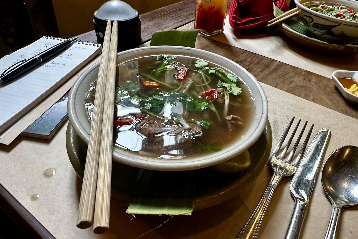 Bowl of herb noodle soup with cilantro and red chilies on a wooden table; chopsticks rest across the bowl beside a notebook and pen.