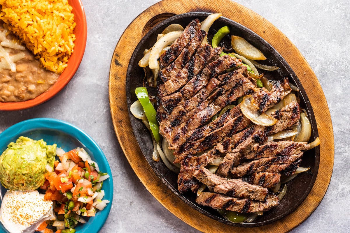 Sizzling beef fajitas with grilled onions and peppers are served with rice, beans, guacamole, and pico de gallo at E Bar, which earned 18 recommendations from Dallas diners. | Photo by E Bar