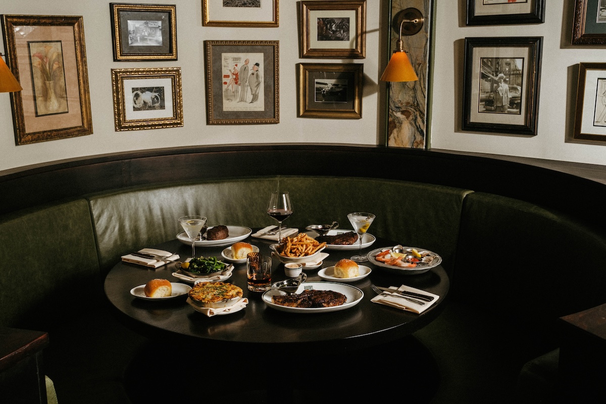 NY_Booths - DiningOut Cozy restaurant booth with a round table loaded with plates of burgers, fries, salad, steak and wine glasses against a wall of framed art.