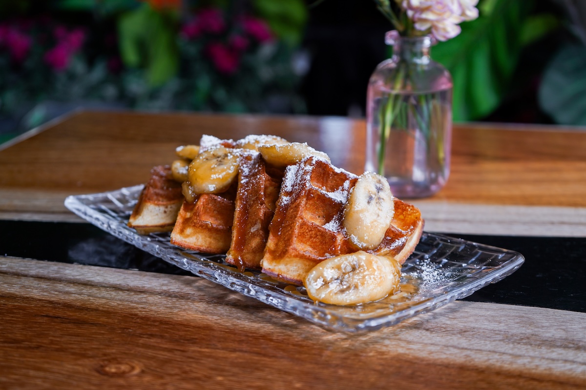 Waffle stack topped with banana slices, dusted with powdered sugar, on a decorative glass platter with syrup drizzle.