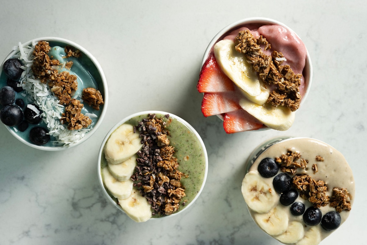 Four smoothie bowls topped with granola, sliced banana, blueberries, and strawberries on a light marble surface (colorful flavors).] ,