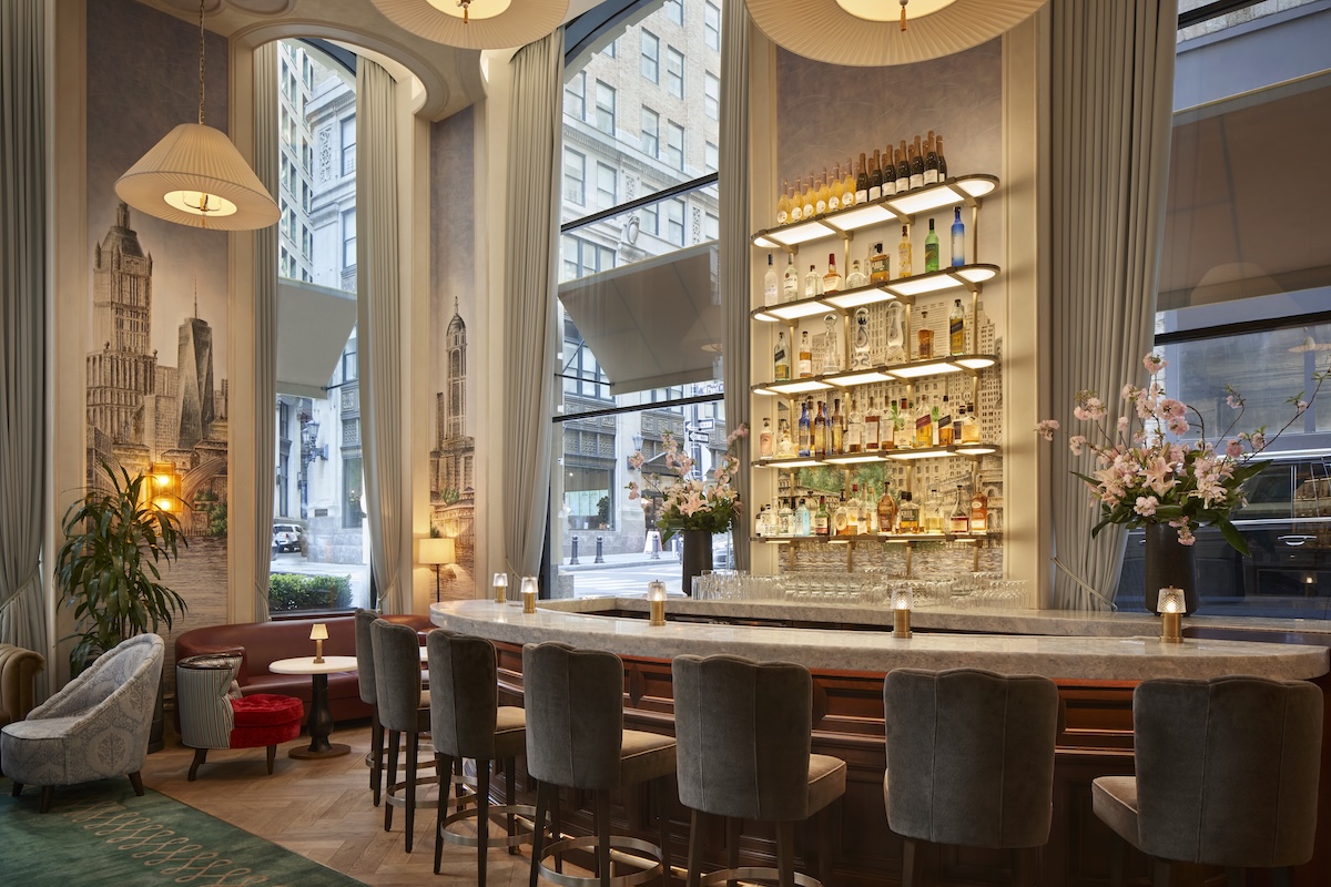 Elegant bar interior with a marble counter, gray upholstered stools, and backlit liquor shelves by tall windows with draped curtains.