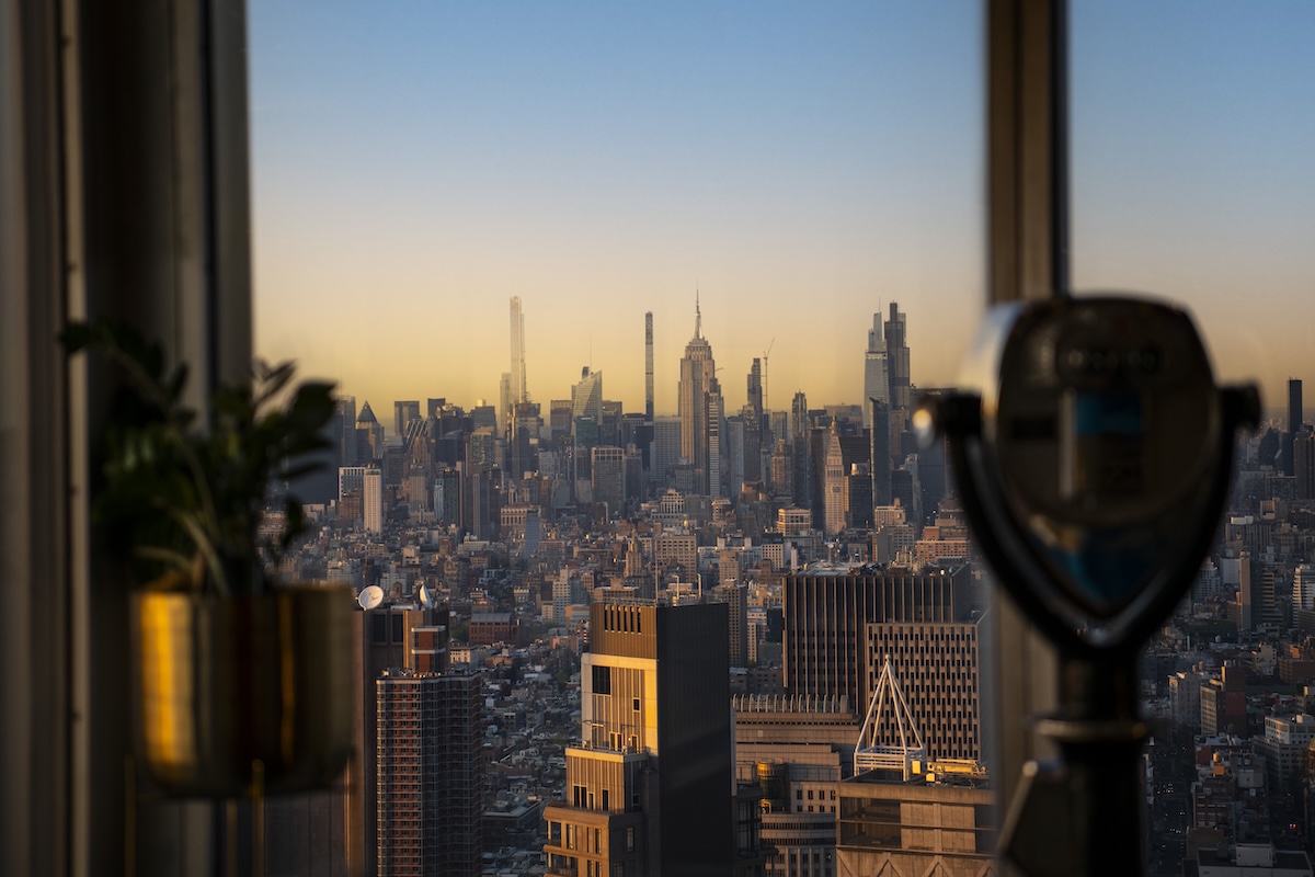 Sunset over a dense city skyline viewed from a high-rise window, with a viewing telescope and potted plant in the foreground.