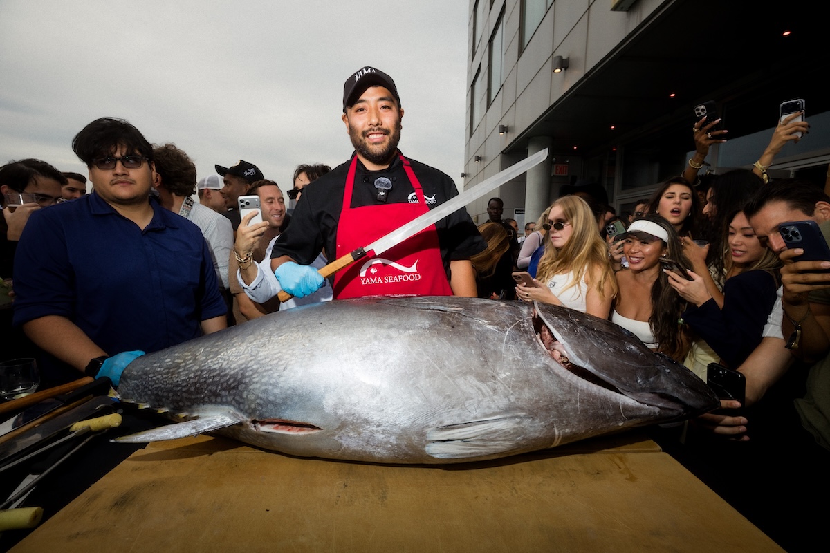 Chef in a red apron and cap stands beside a large swordfish on a table as a crowd of people surrounding him take photos and watch.