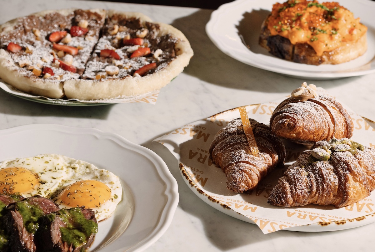 Assorted breakfast pastries on a marble table: powdered-sugar croissants, a chocolate dessert pizza with strawberries, and a plate of sunny-side-up eggs with meat and herbs.