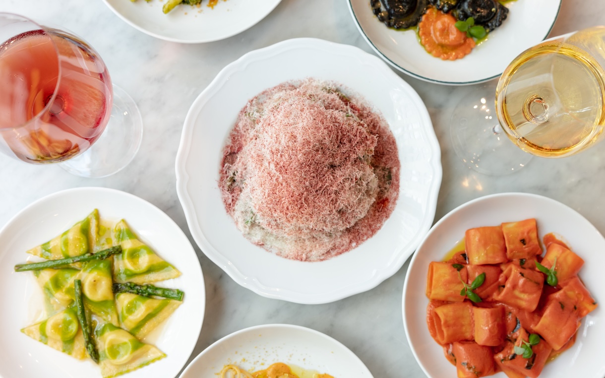 Top-down view of a dinner spread: a central pink fluffy mound in a white bowl, surrounded by plates of pasta with asparagus, cubed melon, and two glasses of wine/rose.