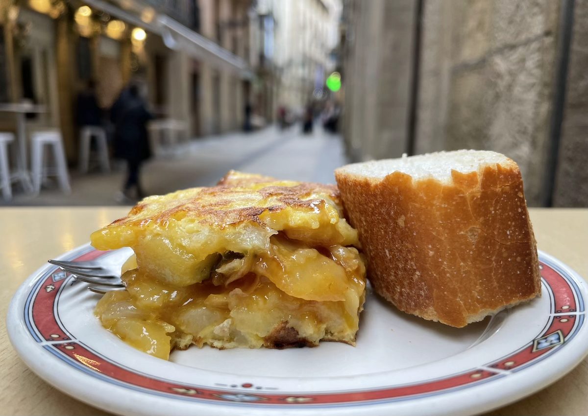 NY_Pintxo_tapa_PhotoBy_DavidFarley - DiningOut Cheesy potato bake on a plate with a crusty bread roll, outdoor cafe setting in the background.