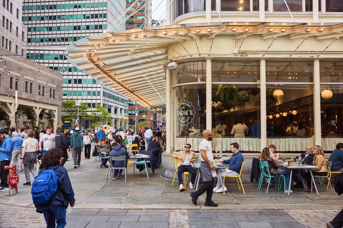 Busy city sidewalk cafe with people dining at colorful chairs outside a restaurant named Sadie’s.