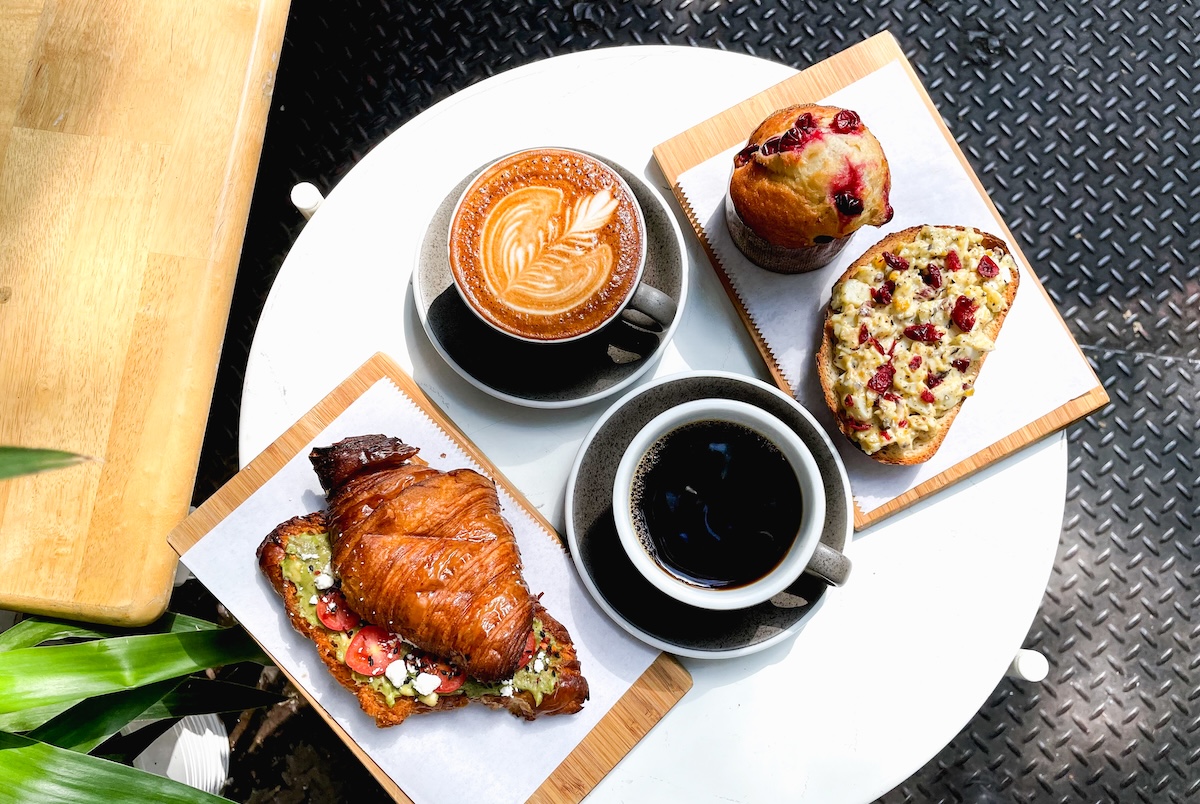 Overhead view of a white round table with two coffee cups (latte with leaf pattern and black coffee) and assorted pastries on wooden boards, at a cafe.