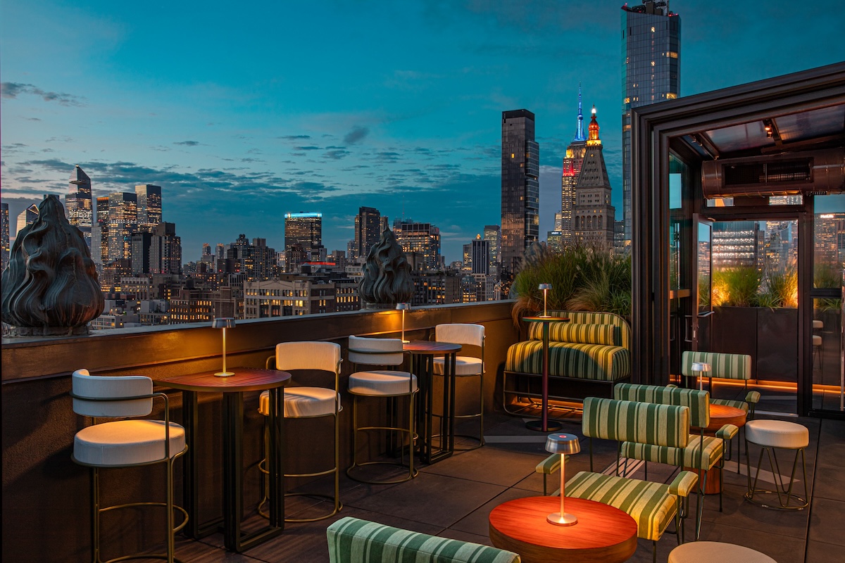Rooftop bar at dusk with city skyline, striped green chairs, and warm table lamps along high stools by a railing.