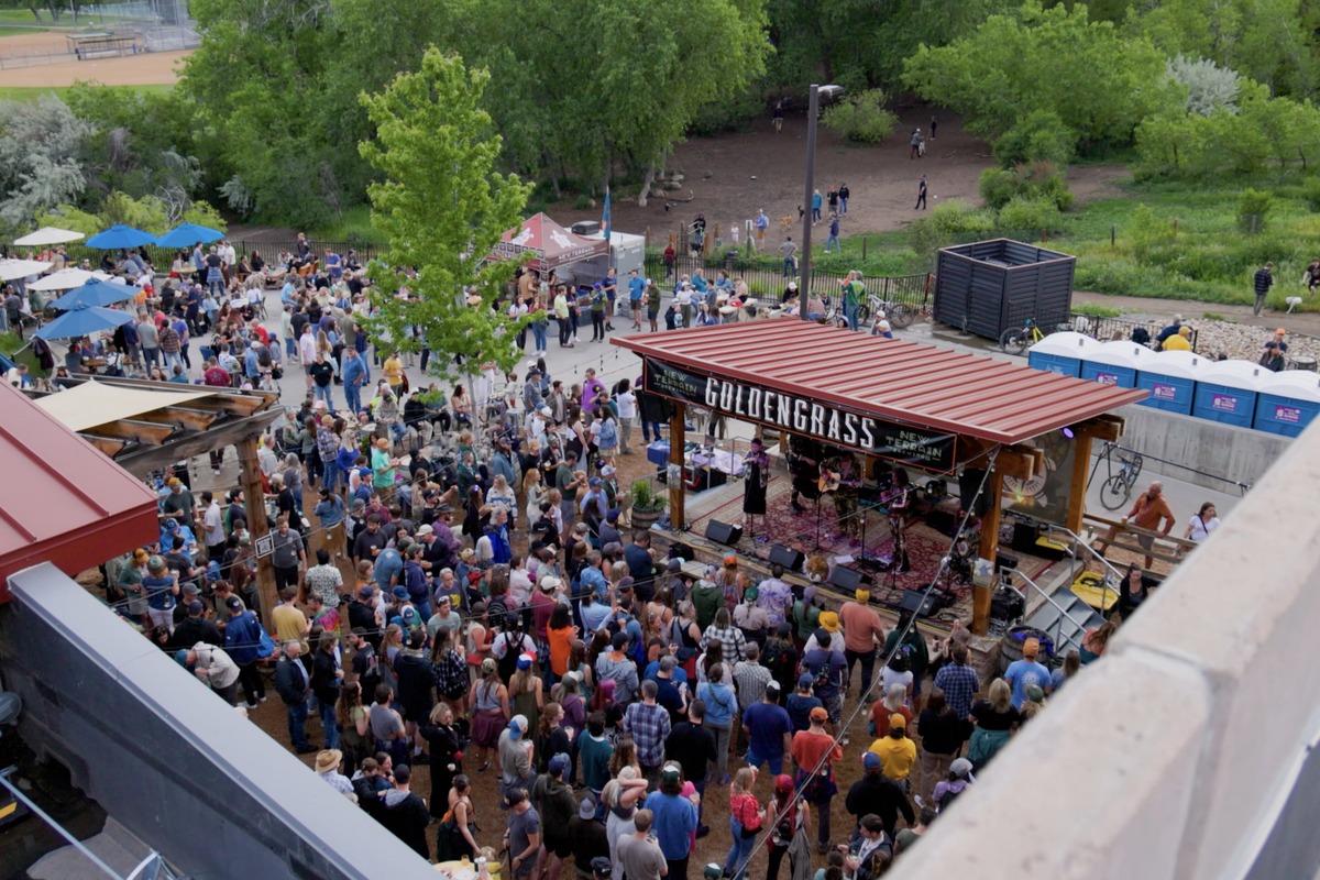 Bird's-eye view of a park festival with a red-roofed stage named GOLDENGRASS, large crowd, and vendor tents amid trees.