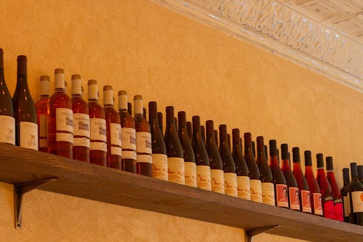 NYC_lai rai wine shelf + Mikey Jackson - DiningOut Row of wine bottles lined up on a wooden shelf against a warm yellow wall.