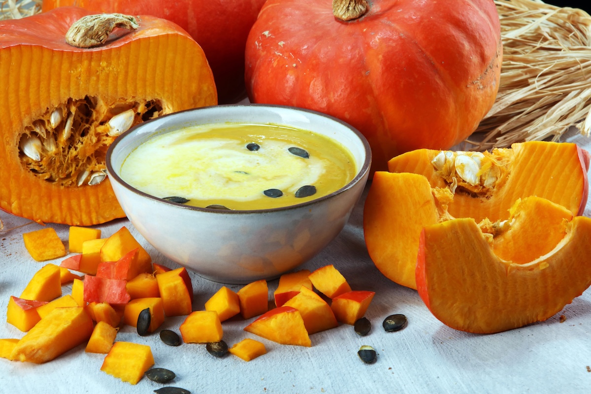 Creamy pumpkin soup in a bowl surrounded by cut pumpkins, cubes of pumpkin flesh, and pumpkin seeds on a light surface.